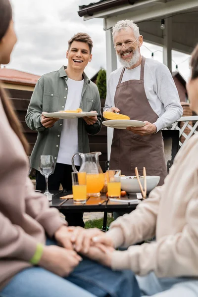 Bearded father and his young adult son holding plates with grilled corn during family bbq party, middle aged man looking at wife and teenage daughter on blurred foreground, backyard — Stock Photo