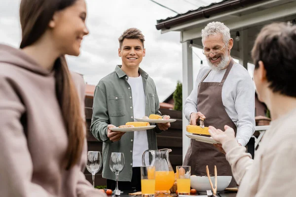 Bearded father and his young adult son holding plates with grilled corn during bbq party, young adult son looking at mother and teenage sister on blurred foreground, backyard — Stock Photo