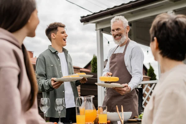 Young adult son smiling near bearded father holding tongs with grilled corn during family bbq party, middle aged man looking at wife and teenage daughter on blurred foreground, backyard — Stock Photo