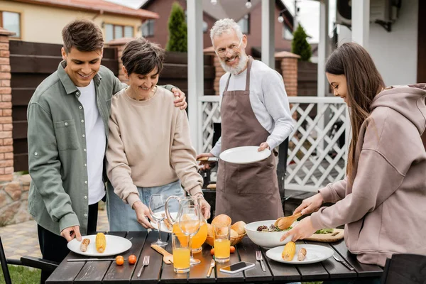 Family bbq party, celebration of parents day, happy young adult son hugging middle aged mother, bearded father in apron looking at teenage daughter, backyard of family house — Stock Photo