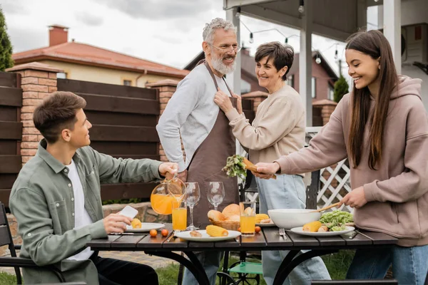 Happy middle aged couple on backyard, joyful teenage girl serving salad near adult brother pouring orange juice, celebrating parents day, love, family grill party, summer — Stock Photo