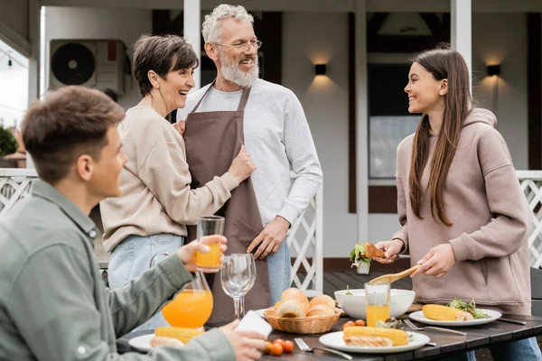 Celebrating parents day, happy middle aged couple looking at joyful teenage girl serving salad near adult brother with glass of orange juice, love, family grill party, summer — Stock Photo