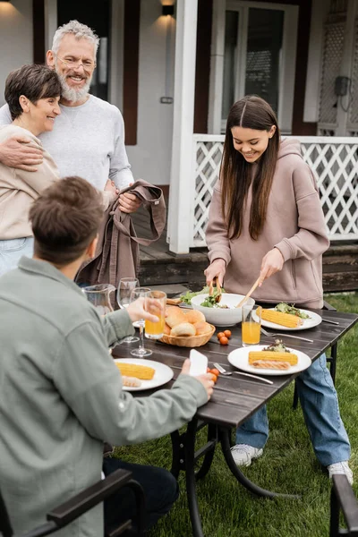 Celebrating parents day, cheerful middle aged couple hugging near joyful teenage girl serving salad next to adult brother with glass of orange juice, love, family grill party, summer, happy marriage — Stock Photo