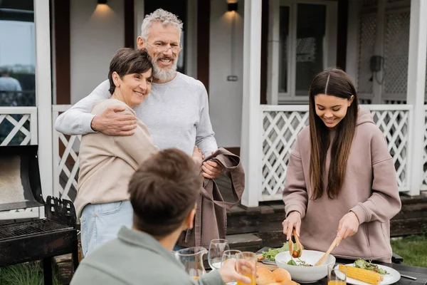 Celebrating parents day, cheerful middle aged couple hugging near joyful teenage daughter serving salad next to adult brother, love, family grill party, summer, happy marriage, summer house backyard — Stock Photo