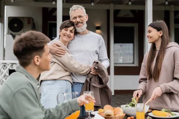 Happy marriage, celebrating parents day, cheerful middle aged couple hugging near joyful teenage daughter mixing salad next to adult brother, love, family grill party, summer,  summer house backyard — Stock Photo