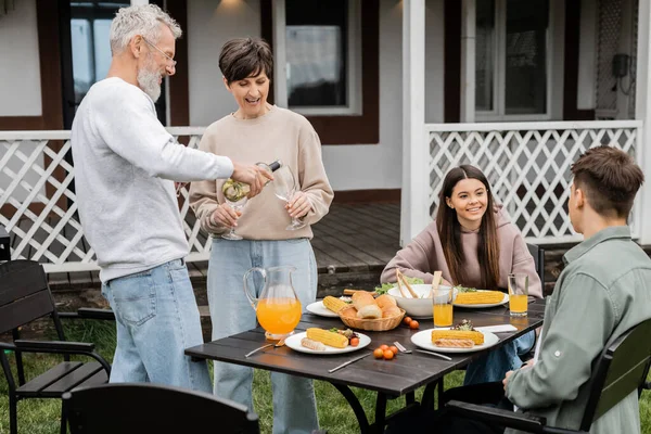 Family bbq party, middle aged man pouring wine into glass of wife near teenage daughter and adult son, grilled food, celebrating parents day on backyard of summer house, june — Stock Photo