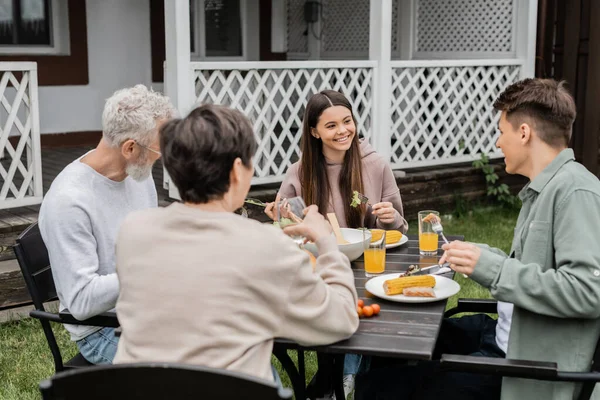 Счастливая девочка-подросток смотрит на брата во время семейного торжества, сидит на заднем дворе дачи, проводит время вместе, ест гриль-барбекю, радуется родительскому дню — стоковое фото