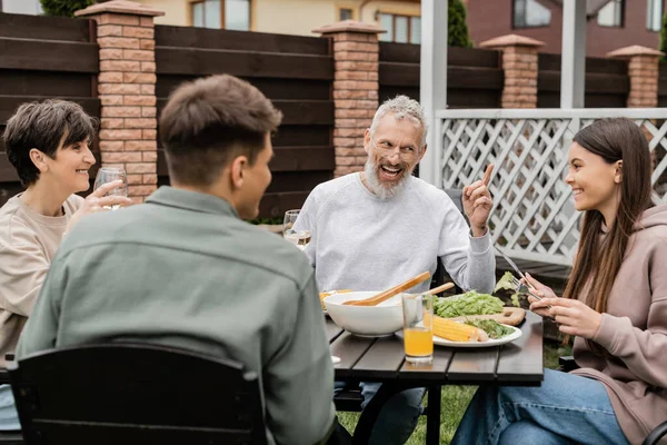 Cheerful middle aged man looking at teenage daughter and gesturing during bbq party, sitting on backyard of summer house, spending time together, eating grilled bbq food, happy parents day concept — Stock Photo