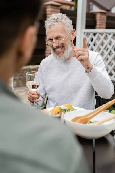 Cheerful middle aged man looking son and gesturing during bbq party, blurred foreground, sitting on backyard of summer house, spending time together, eating grilled bbq food, parents day concept — Stock Photo