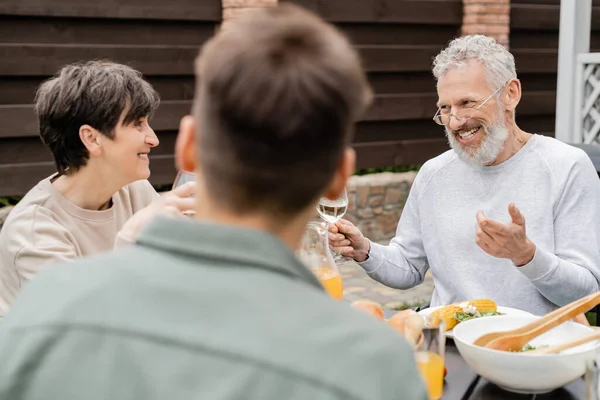 Cheerful middle aged man looking wife near adult son and gesturing during bbq party, blurred foreground, backyard of summer house, spending time together, eating grilled bbq food, parents day concept — Stock Photo