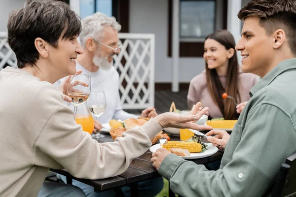 Happy middle aged mother holding glass of wine and talking with adult son during bbq family party, eating grilled food, father and daughter on blurred background, backyard of summer house — Stock Photo