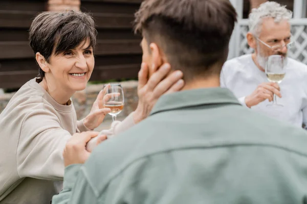 Sorrindo mãe de meia idade segurando copo de vinho e tocando o filho adulto borrado perto do marido no fundo durante a celebração do dia dos pais no quintal, amor familiar e conceito de unidade — Fotografia de Stock