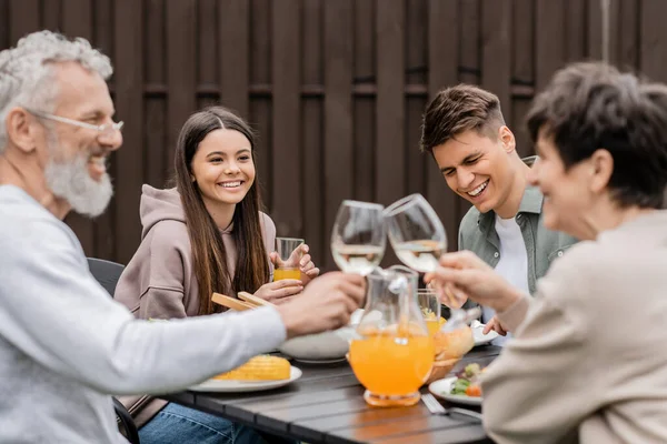 Smiling siblings sitting near tasty bbq food and blurred middle aged parents toasting with wine during parents day celebration at backyard, family love and unity concept, tradition and celebration — Stock Photo