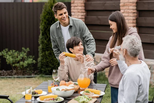 Smiling siblings talking to middle aged parents with glasses of wine near summer food during family barbeque and parents day celebration at backyard, family love and unity concept — Stock Photo