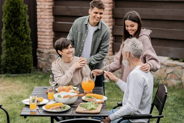 Smiling siblings hugging middle aged parents and spending time together near summer food during bbq party and parents day celebration at backyard, family love and unity concept — Stock Photo