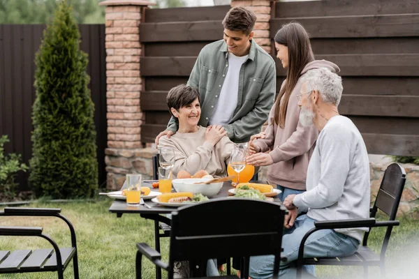 Smiling middle aged woman looking at husband near positive children and summer food during bbq party and parents day celebration at backyard, family love and unity concept, special occasion — Stock Photo