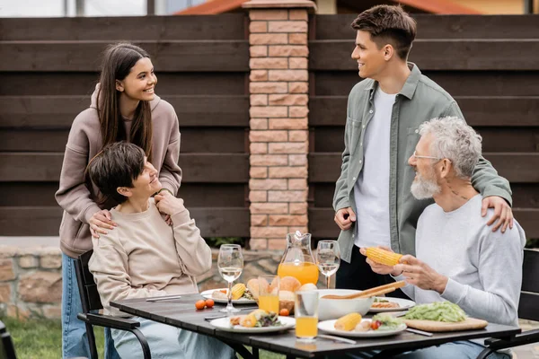 Positive family with kids talking while spending time together near summer barbeque food during parents day celebration at backyard in june, family love and unity concept, tradition and celebration — Stock Photo