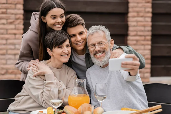 Sourire papa d'âge moyen prendre selfie sur smartphone avec la famille et les enfants près de savoureux repas d'été lors de la fête barbecue et fête des parents à l'arrière-cour, l'amour de la famille et concept d'unité — Photo de stock
