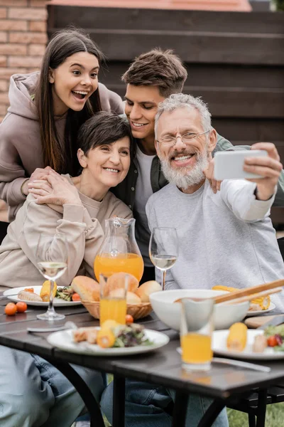 Positiver reifer Vater macht Selfie auf Smartphone mit Frau und Kindern in der Nähe von verschwommenem Sommeressen bei Grillparty und Elterntagsfeier im Hinterhof, glückliches Elterntagskonzept — Stockfoto