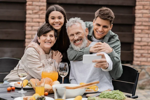 Lächelnde Eltern mittleren Alters, die ihre Kinder umarmen und in die Kamera schauen, während sie während der Grillparty und der Elterntagsfeier im Hinterhof im Juni ein Selfie mit dem Smartphone machen. — Stockfoto