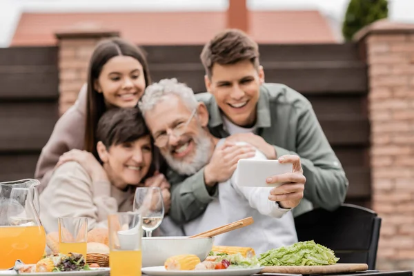 Smartphone in hand of blurred mature man taking selfie with family and kids near summer food during bbq party and parents day celebration at backyard in june, happy parents day concept — Stock Photo