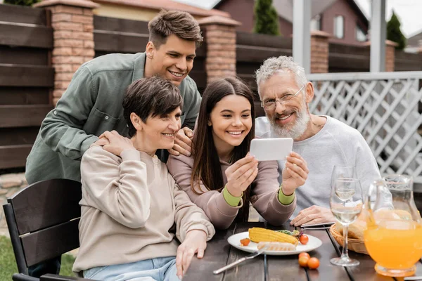 Smiling teen girl using smartphone near middle aged parents and brother during barbeque party with summer food and parents day celebration at backyard in june, happy parents day concept — Stock Photo