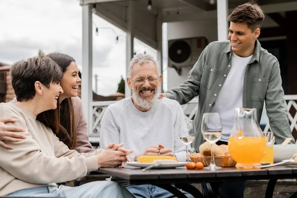 Positive woman and kids looking at laughing middle aged husband near summer food during barbeque party and parents day celebration at backyard in june, happy parents day concept — Stock Photo