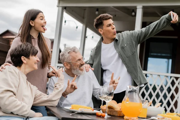 Young man sticking out tongue while taking selfie with sister and cheerful middle aged parents during bbq party and parents day celebration at backyard in june, happy parents day concept — Stock Photo