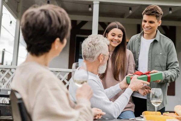 Smiling siblings giving gift box to blurred middle aged parents with wine near food during barbeque party and parents day celebration at backyard in june, celebrating parenthood day concept — Stock Photo
