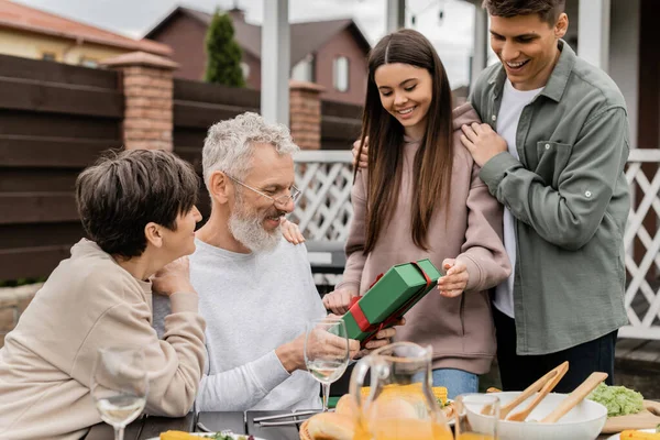 Smiling siblings giving present box to middle aged parents near summer food during barbeque party and parents day celebration at backyard in june, celebrating parenthood day concept — Stock Photo
