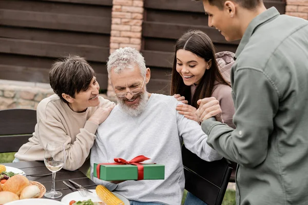 Smiling middle aged man holding gift box near children and wife during barbeque party with summer food and parents day celebration at backyard in june, celebrating parenthood day concept — Stock Photo
