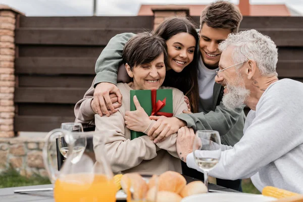 Cheerful kids hugging positive mother holding gift box near father and summer food during barbeque party and parents day celebration at backyard in june, celebrating parenthood day concept — Stock Photo