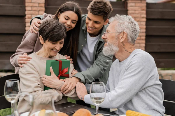 Cheerful middle aged man and children hugging woman with gift box near summer food during barbeque party and parents day celebration at backyard in june, celebrating parenthood day concept — Stock Photo