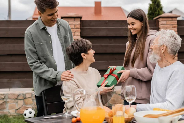 Smiling teenage daughter giving gift box to middle aged mother near brother and father during parents day celebration and barbeque party at backyard in june, celebrating parenthood day concept — Stock Photo
