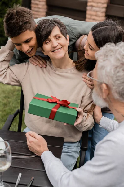 Femme d'âge moyen souriante tenant une boîte-cadeau et embrassant les enfants près du mari flou pendant le pique-nique et la célébration de la journée des parents à l'arrière-cour en juin, célébrant le concept de jour de la parentalité — Photo de stock