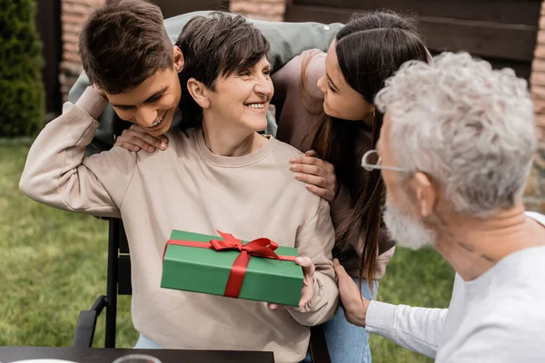 Positive children hugging middle aged mother holding gift box near blurred husband while spending time during parents day celebration at backyard in june, cherishing family bonds concept — Stock Photo