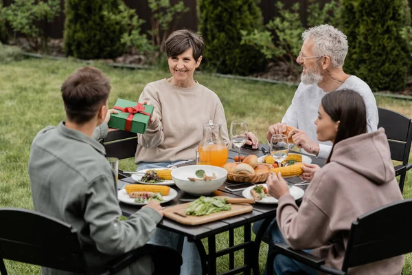 Smiling middle aged mother taking gift box from young son near family during summer food and bbq party during parents day celebration at backyard in june, cherishing family bonds concept — Stock Photo