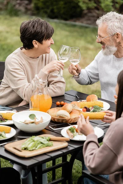 Smiling middle aged wife and husband toasting glasses of wine near summer food and teenage daughter during bbq party and parents day celebration at backyard, cherishing family bonds concept — Stock Photo
