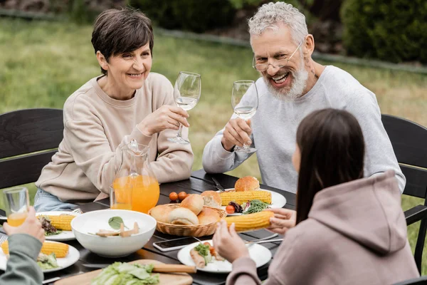 Cheerful middle aged parents holding glasses of wine near summer food and children during barbeque party and parents day celebration at backyard in june, cherishing family bonds concept — Stock Photo
