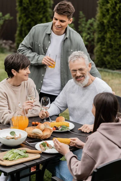 Smiling kids talking to middle aged parents near tasty summer food and parents day celebration during barbeque party at backyard in june, cherishing family bonds concept, special occasion — Stock Photo
