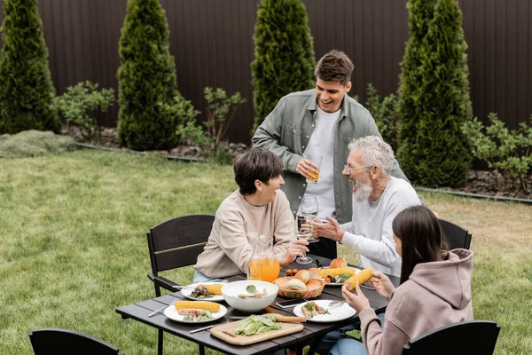 Excited and cheerful middle aged parents toasting with wine glasses near children and summer food during bbq party at backyard, cherishing family bonds concept, spending time together — Stock Photo