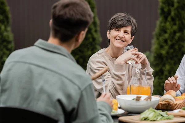 Smiling middle aged woman holding glass of wine and looking at blurred young son near husband and summer food during parents day celebration at backyard, cherishing family bonds concept — Stock Photo