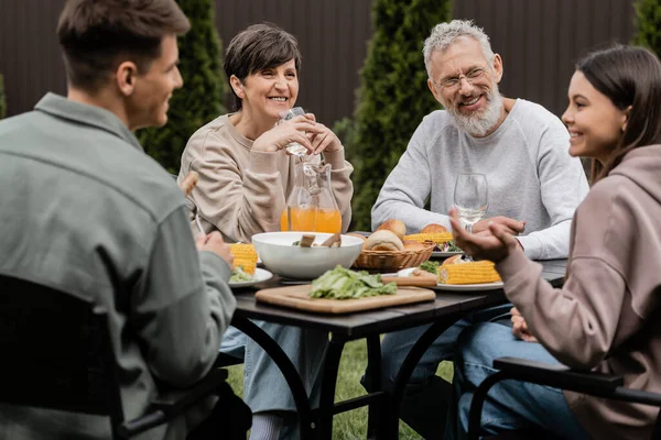 Padres positivos de mediana edad mirando a los niños borrosos cerca de la comida de verano durante la fiesta de barbacoa y la celebración del día de los padres en el patio trasero en junio, apreciando el concepto de lazos familiares - foto de stock