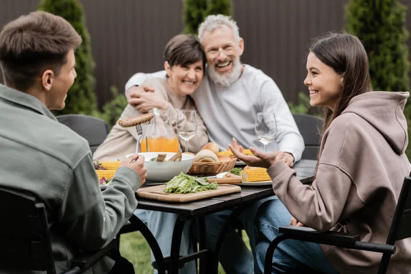 Усміхнені діти розмовляють один з одним, сидячи біля літньої їжі та розмитих батьків середнього віку на фоні під час святкування дня батьків на фоні, плекаючи концепцію сімейних зв'язків — стокове фото