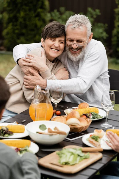 Cheerful middle aged man hugging wife while celebrating parents day near blurred kids and tasty summer food during bbq party at backyard in june, special day for parents concept — Stock Photo