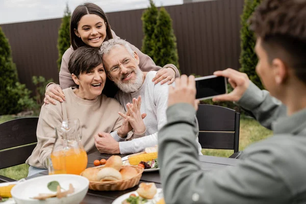 Lächelndes Teenager-Mädchen umarmt Eltern mittleren Alters in der Nähe verschwommenen Bruder, der Foto auf dem Smartphone in der Nähe von BBQ-Essen während der Eltern-Tag-Feier im Hinterhof, besonderen Tag für Eltern Konzept — Stockfoto