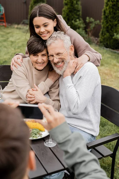Smiling teenage girl hugging mature parents while blurred brother taking photo on smartphone during barbeque party and parents day celebration at backyard, special day for parents concept — Stock Photo
