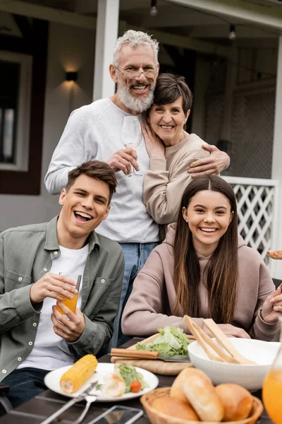 Cheerful mature parents hugging and looking at camera near children and summer food while celebrating parents day at backyard in june, special day for parents concept, tradition and celebration — Stock Photo