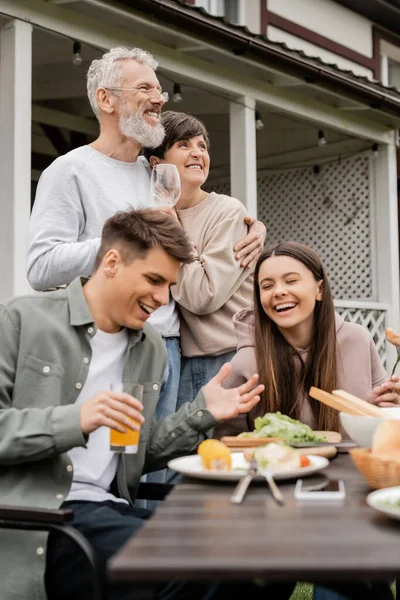 Cheerful middle aged parents hugging while standing near kids and bbq food during parents day celebration and party at backyard in june, special day for parents concept, tradition and celebration — Stock Photo