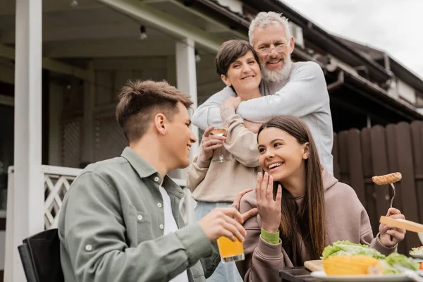 Smiling teenage girl talking to brother while parents hugging during barbeque party and parents day celebration at backyard in june, special day for parents concept, tradition and celebration — Stock Photo